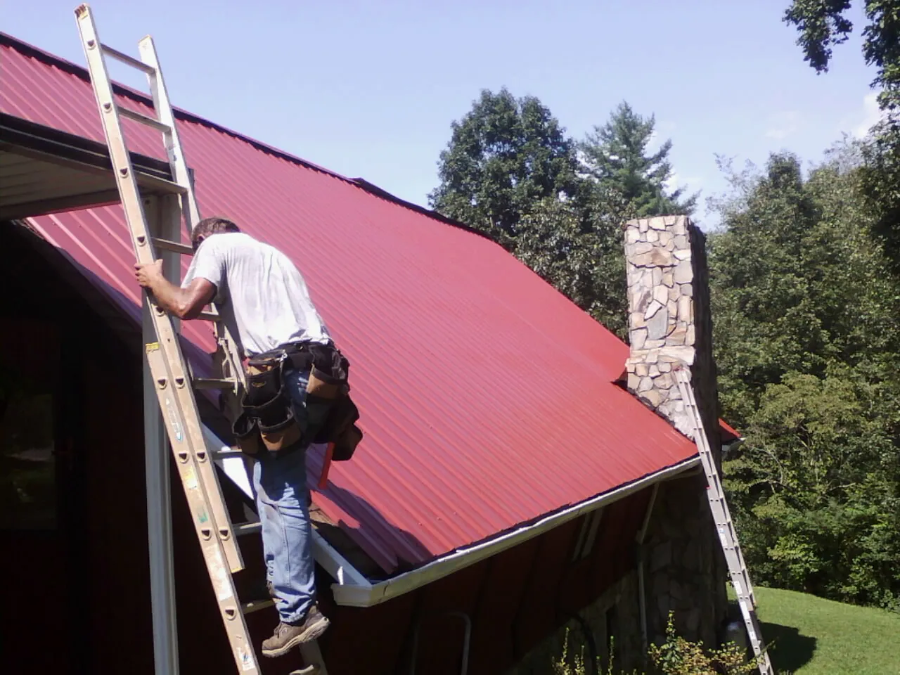 Roofer on a ladder beside a red metal roof and stone chimney — Soffit & Fascia Repair by Roofing Brothers in New Brighton